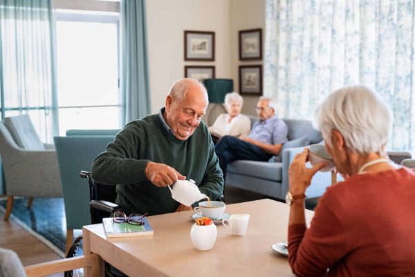 Smiling senior man pouring tea in cup from teapot in care facility with wife sitting at table in the common area. Elderly man in care centre sitting on wheelchair and serving tea. Serene disabled senior man enjoying tea time with friends during a visit at nursing home.